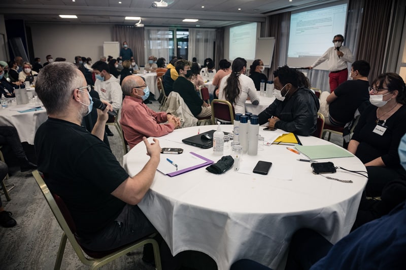 Conférence Citoyenne sur l'aide, l'entraide et les solidarités, 2021 - Réunion plénière : vue d'ensemble de la salle et des participants installés autour de plusieurs tables rondes
